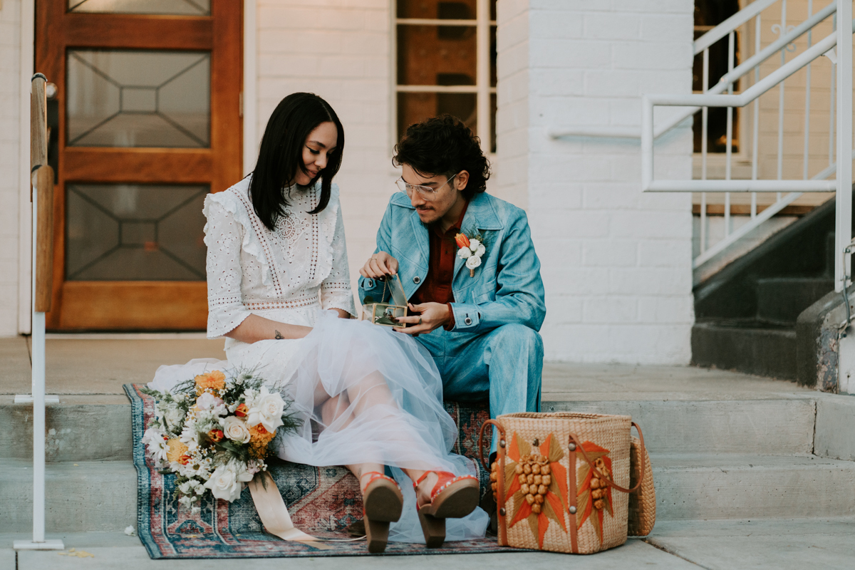 bride and groom dressed in vintage clothing sit on cement stairs in front of a decorative door chatting and eating from a picnic basket.
