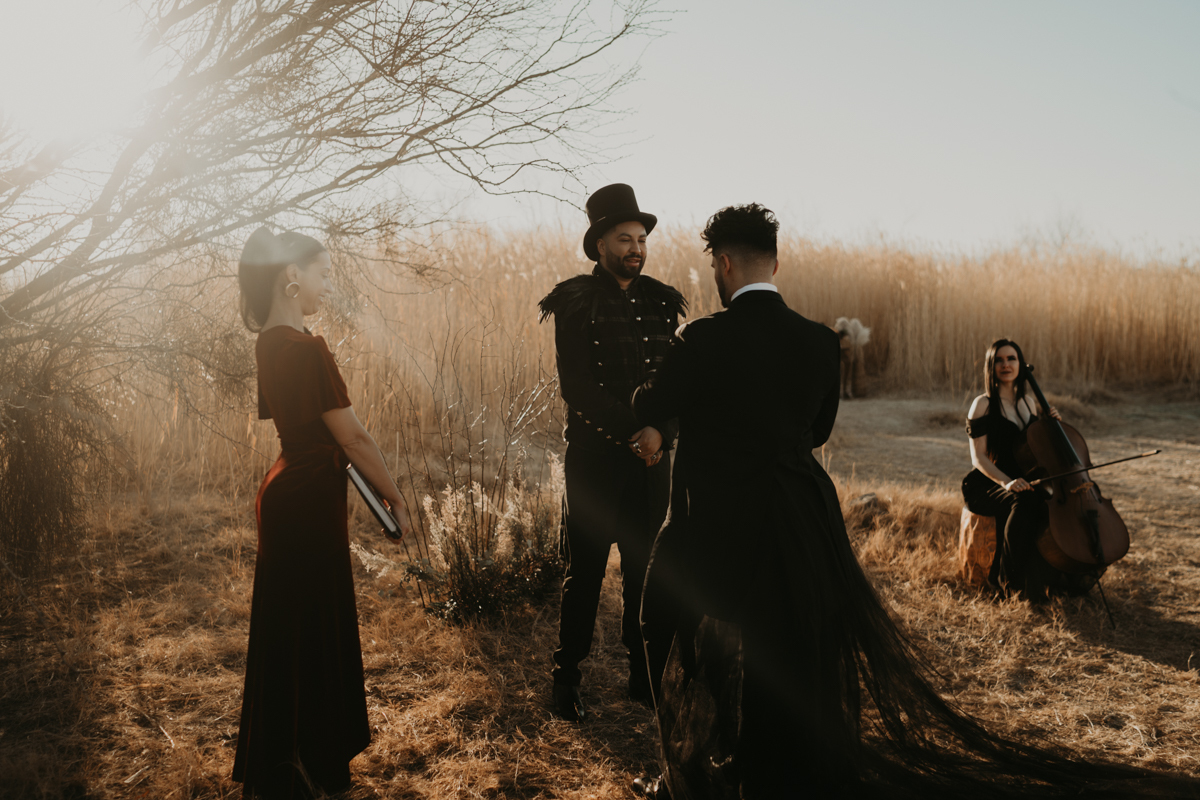 two gothic grooms exchange vows in a wheat field as female officiant and female violinist look on.