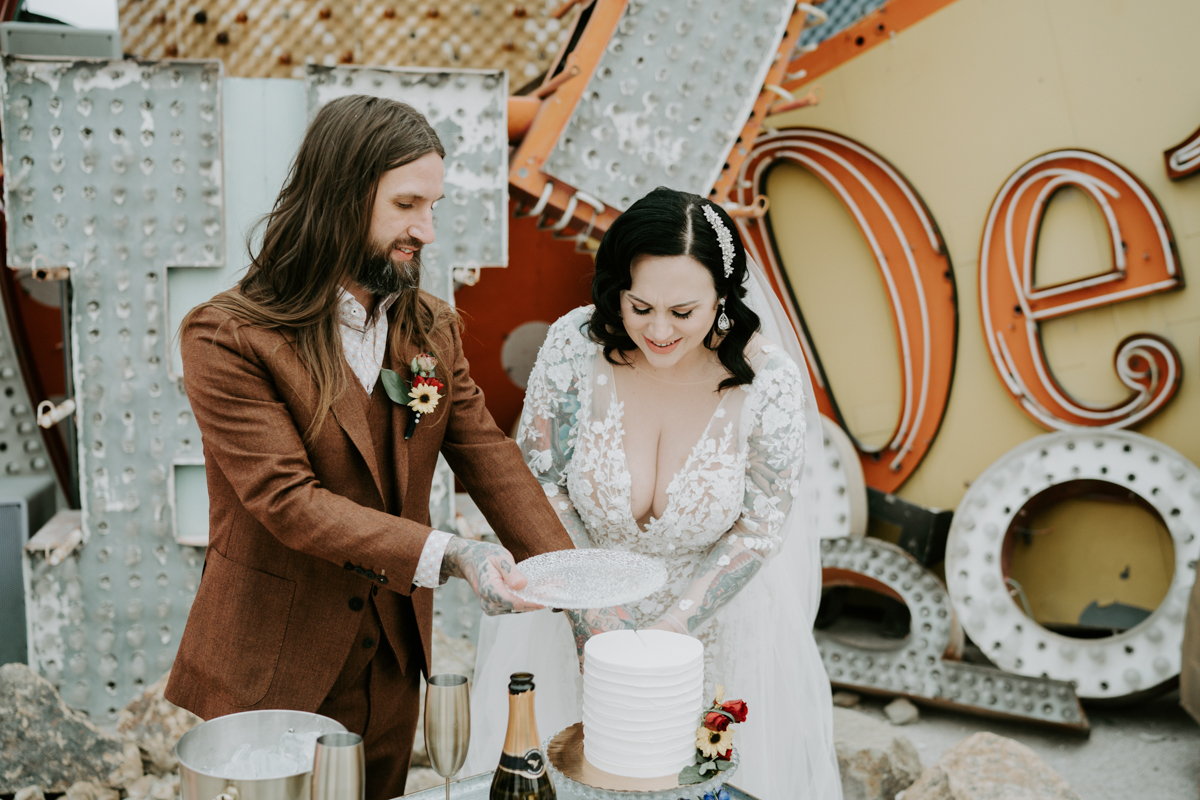 a groom with long hair and a vintage suit holds a plate out as his bride cuts the cake. they’re standing in front of old vintage neon signs.