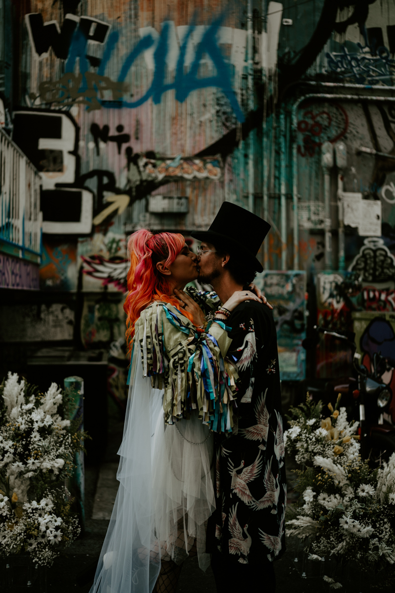 a bride with brightly colored hair kisses her groom wearing a top hat. they stand in front of a building with a lot of graffiti.