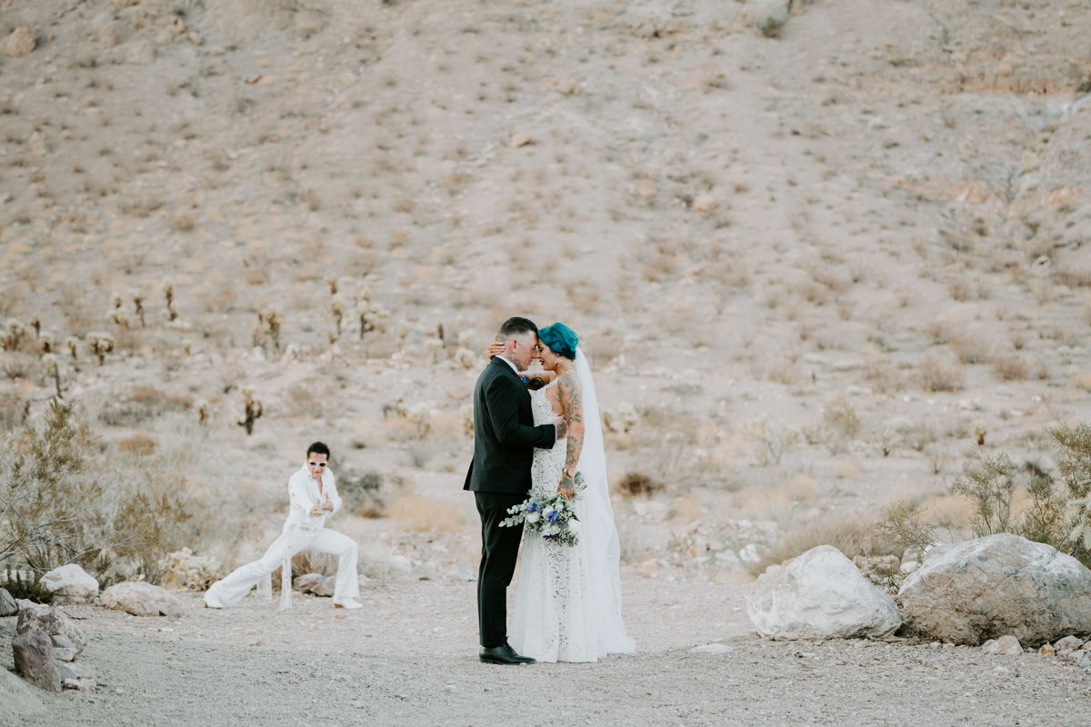 A groom and bride press their foreheads together and embrace in the desert with an Elvis impersonator posing in the background.
