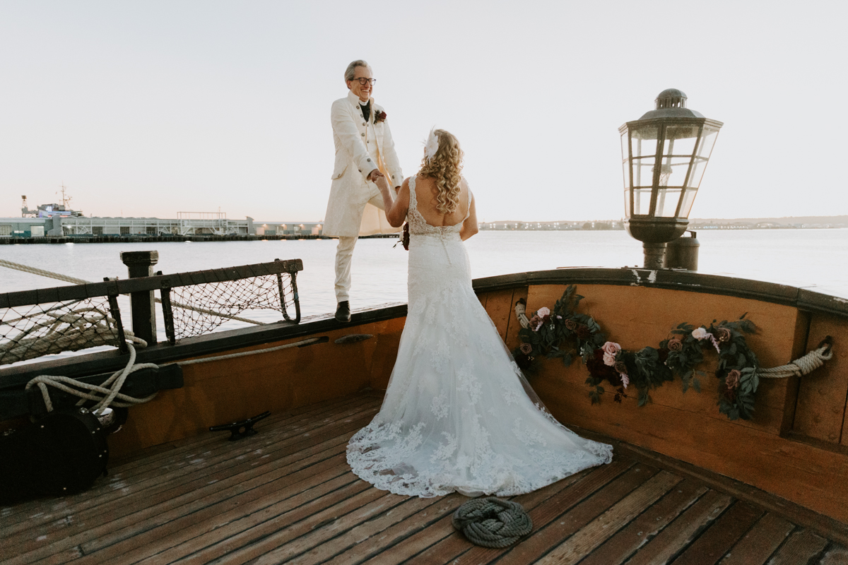 Groom wearing 18th century style white suit stands on the side of a boat and holds his bride's hand and smiles down on her. Her back is to the camera and she's wearing a long white dress.