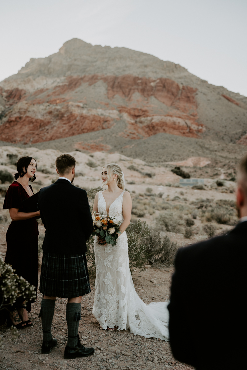 a groom and bride stand in the desert as their female officiant speaks. an onlooking guest is blurred in the foreground.