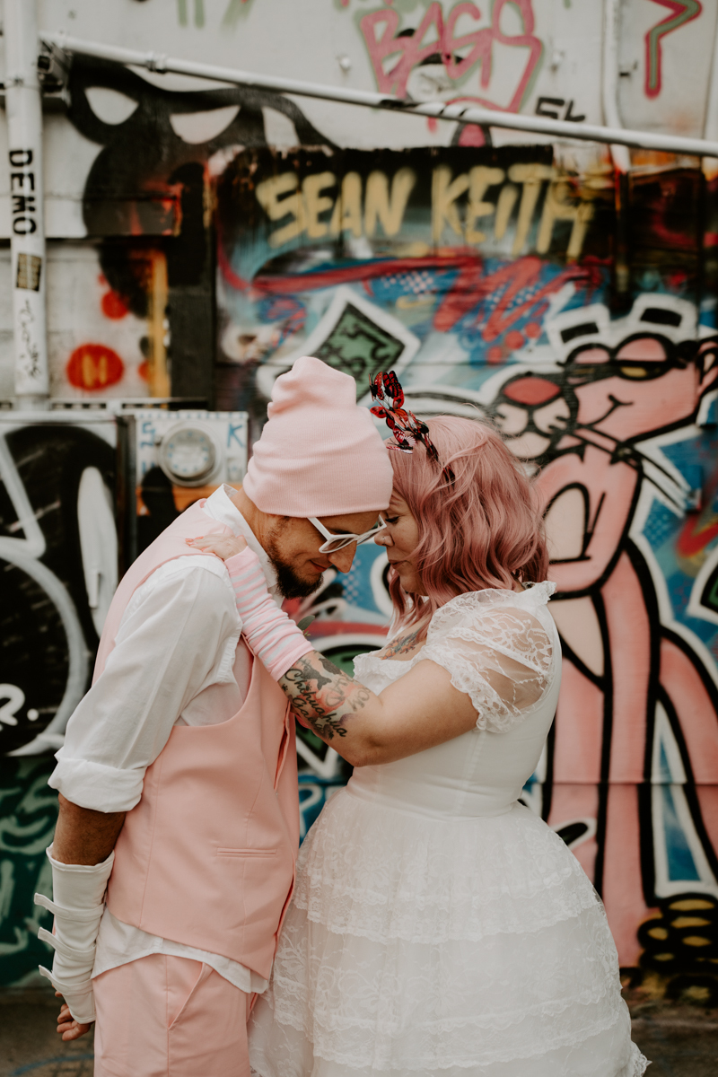groom in pink vest and beanie leans into his bride with pink hair and pink fingerless loves.
