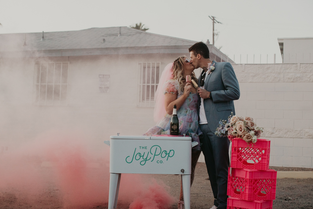 a bride in brightly colored dress kisses her groom as they both hold popsicles. they stand behind a small old fashioned cooler that says the joy of pop. the bride’s bouquet rests on a stack of hot pink milk crates.