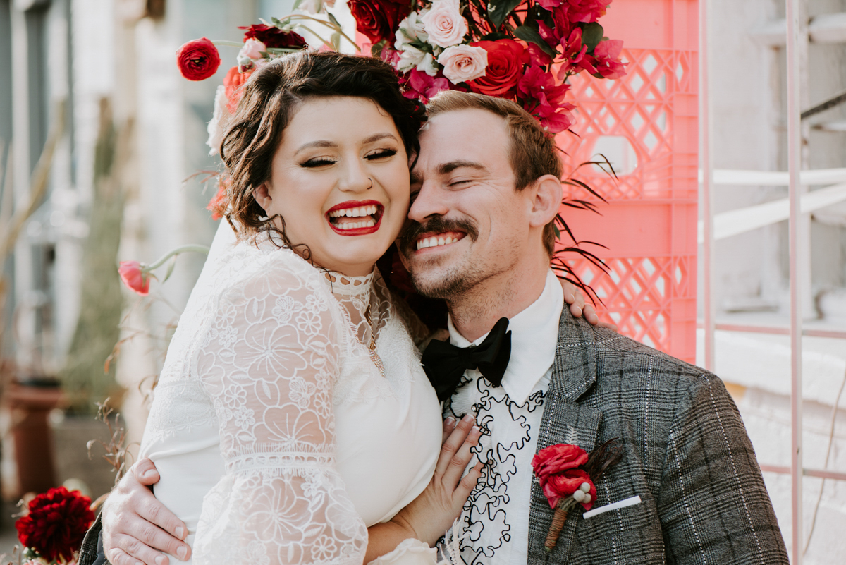a bride and groom embrace and laugh while seated. the groom is wearing a bow tie and ruffle shirt.