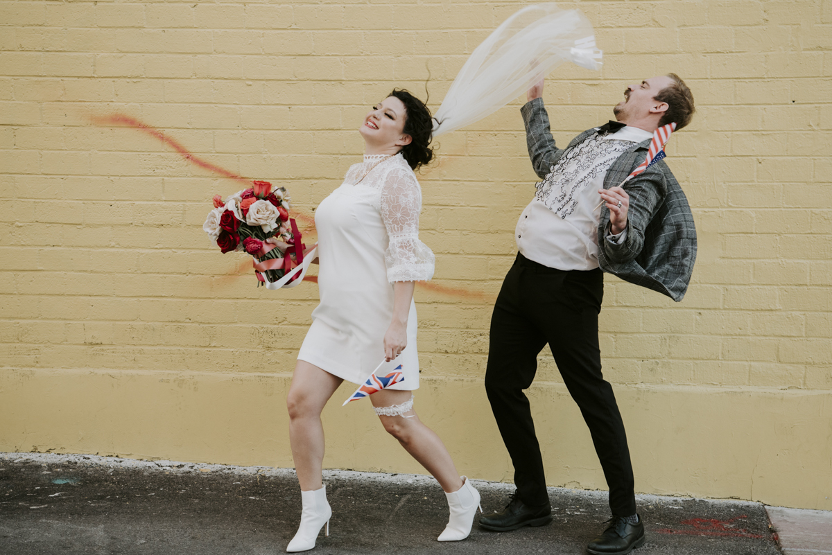 Bride and groom in retro clothing jumping in front of yellow brick wall. Bride has a little UK flag and groom has US flag.