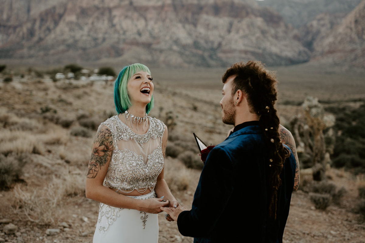 a laughing blue haired bride and groom hold hands and exchange vows as their officiant looks on.