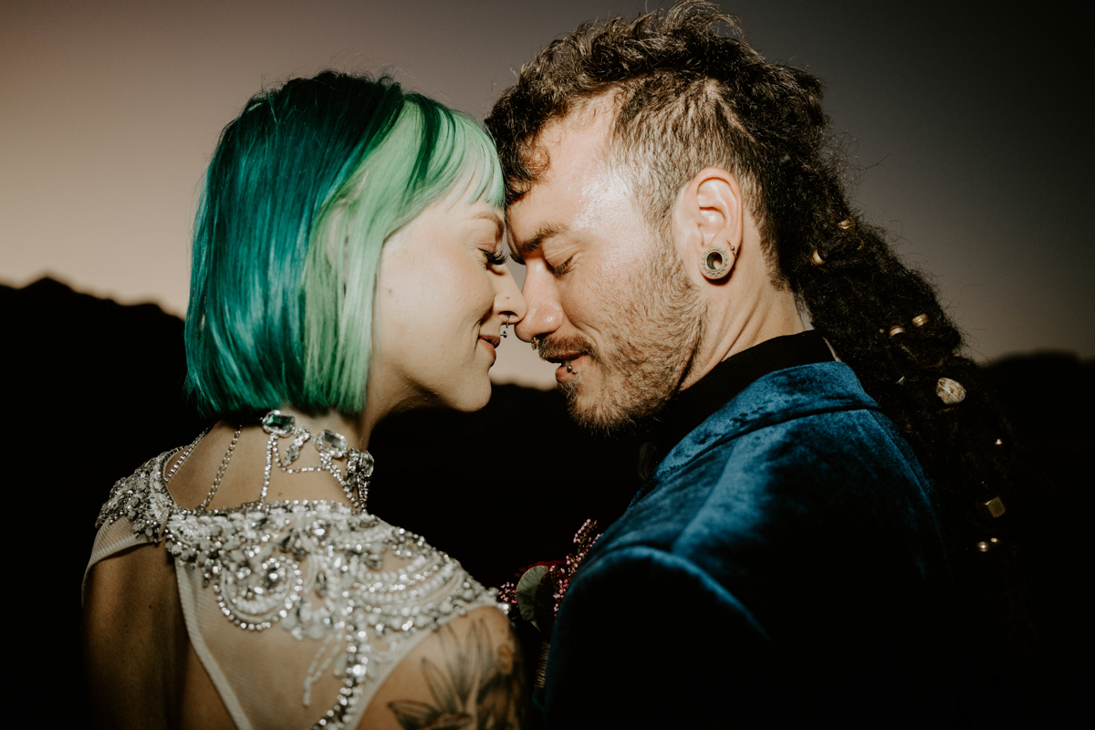 a bride with blue hair presses her forehead to her groom’s forehead. he’s wearing a blue velvet suit.