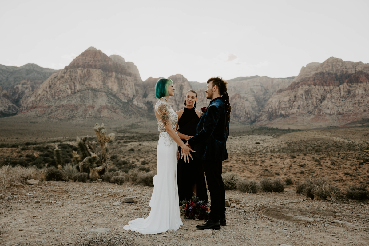 A bride with blue hair stands with her groom and a female officiant in a Vegas desert with mountains in the background.